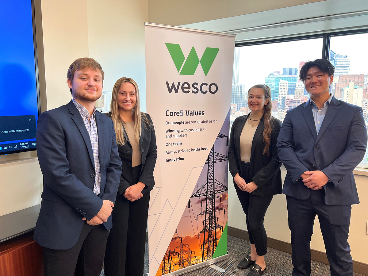 Four students in business attire stand grouped around a Wesco sign. 
