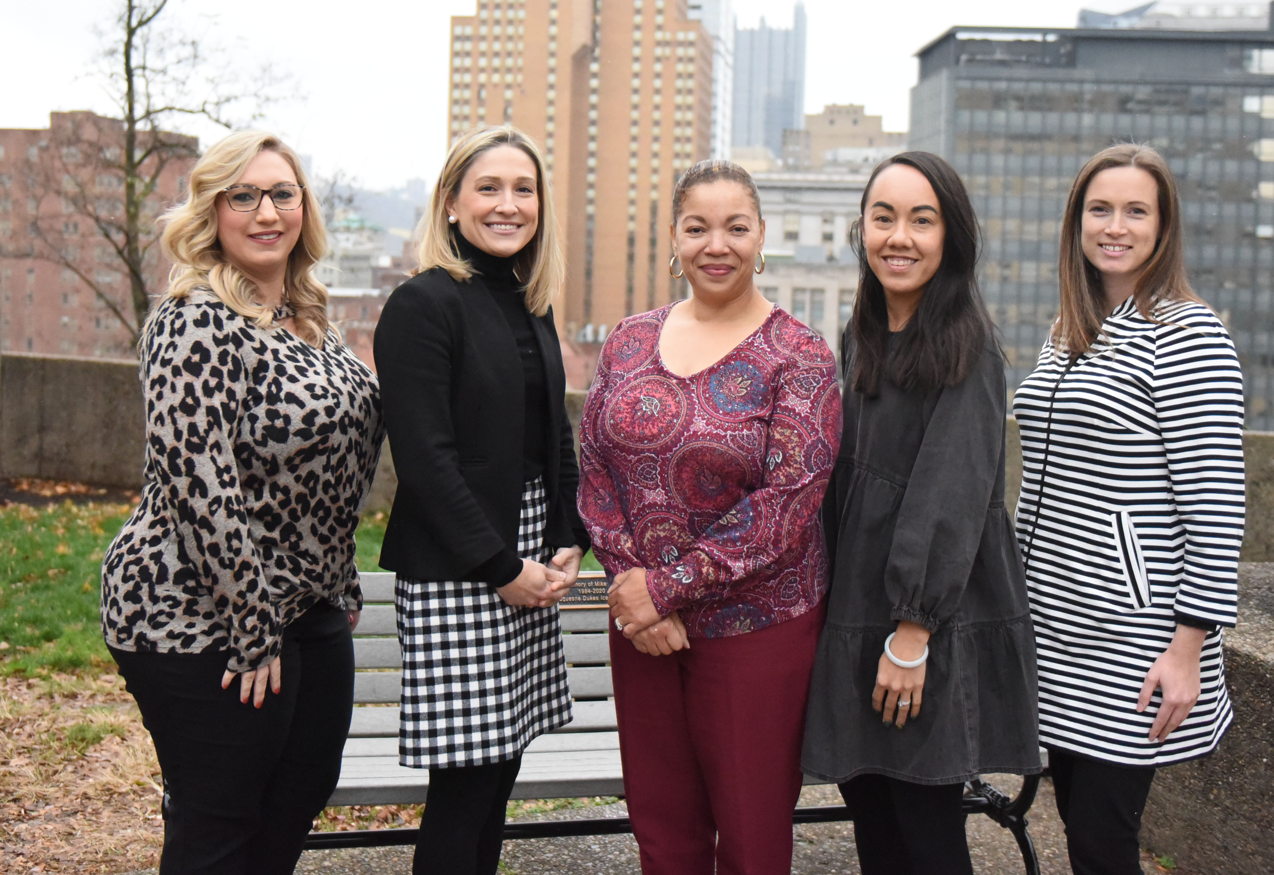 Five women in business attire stand in a row outside with downtown Pittsburgh behind them. 