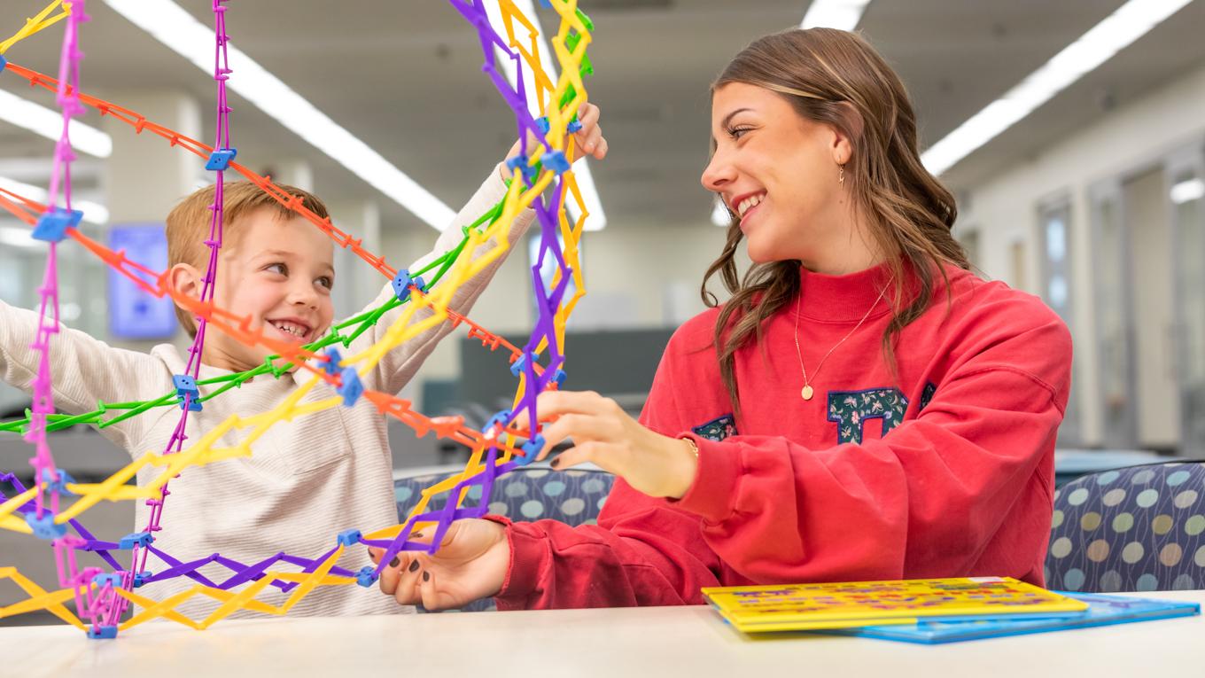 Young learner playing with geometric colorful manipulative looking and smiling at Duquesne Early Childhood Eduation student in library