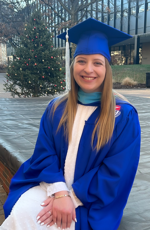 MAT Early Childhood Education graduate Samantha Szewczyk with graduation regalia sitting on the edge of fountain outside on campus with Christmas tree in background