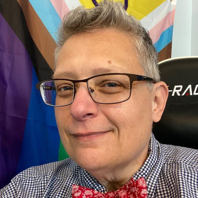 person wearing bowtie and glasses sits in front of a Pride flag