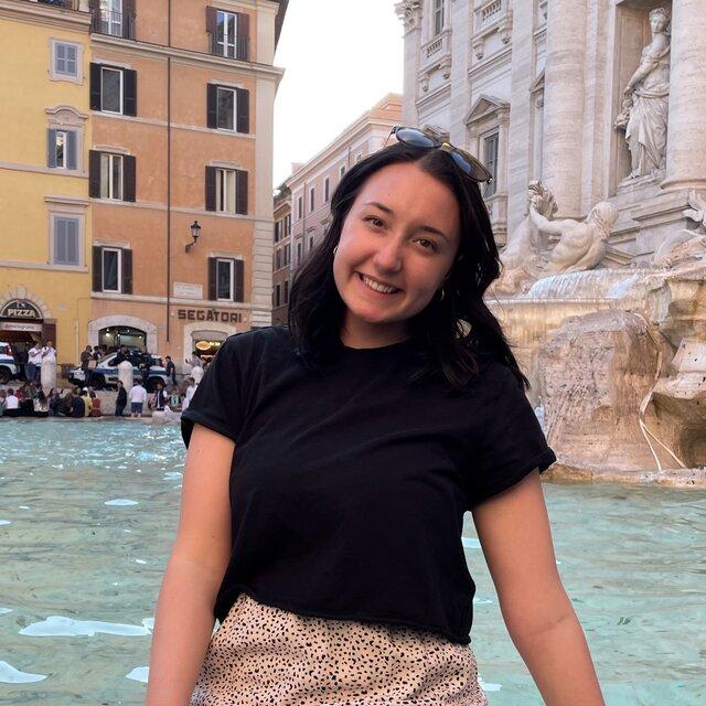 Jessica smiles sitting in front of the Trevi Fountain in Rome in a black shirt.