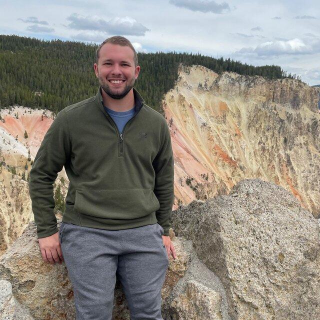 Nathan stands in front of a colorful mountain range in a green sweatshirt.